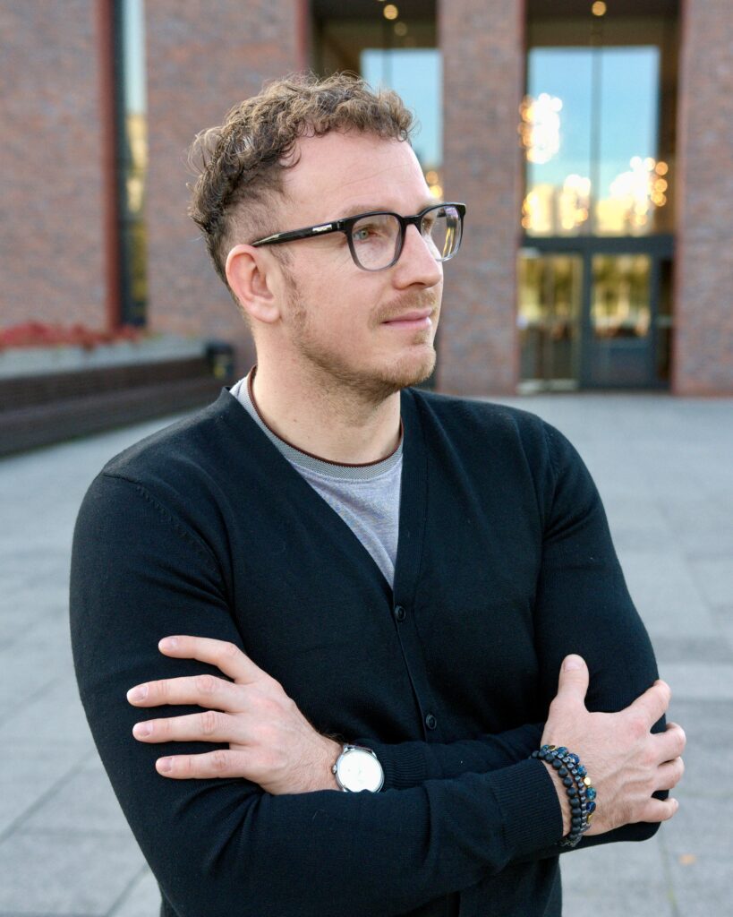 Daniel Nadolski w ciemnym kardiganie na tle architektury